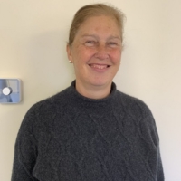 A woman with light brown hair pulled back smiles, wearing a dark jumper, standing against a cream wall.