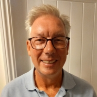 Smiling older man with glasses and grey hair, wearing a light blue polo shirt, standing indoors.