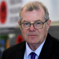 An older man with grey hair and glasses wearing a suit and tie, looking at the camera.
