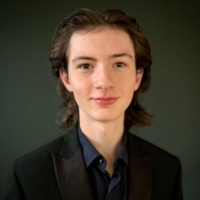 A young person with medium-length brown hair, wearing a dark blazer and shirt, smiles at the camera.
