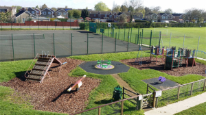 Norwood-Playing-Field--2 A playground with slides, climbing frames, a roundabout, and a fenced tennis court in the background.