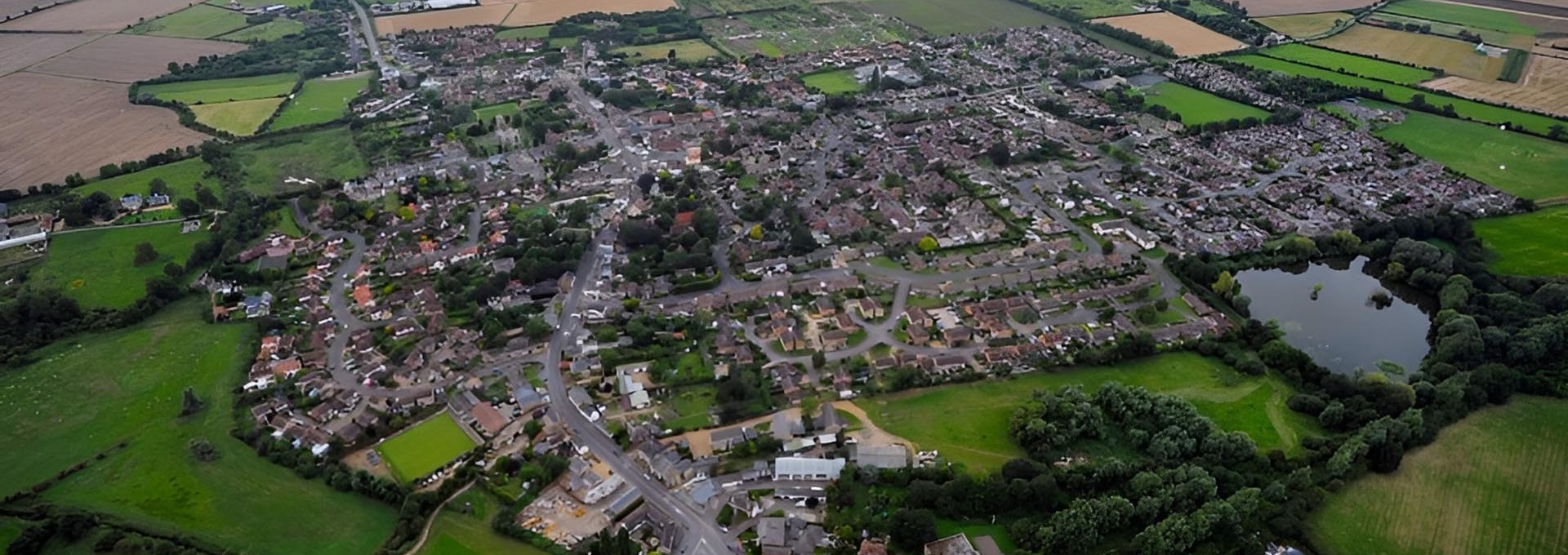 Aerial view of a village with houses, roads, trees, fields, and a small pond.