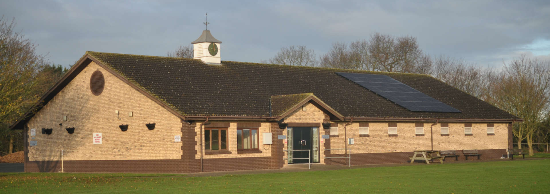 Single-storey brick community building with a clock tower and solar panels, beside a grassy field.